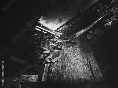 Holzschuppen mit bedrohlich wirkender Axt und großem Holzblock mit viel gestapeltem Feuerholz im Hintergrund. Lichteinfall im Holzschuppen in düsterem monochrom schwarzweiss.
