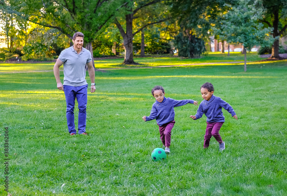 Caucasian father playing soccer with his four year old African-American twin boys in public park Caucasian father playing soccer with his four year old African-American twin boys in public park