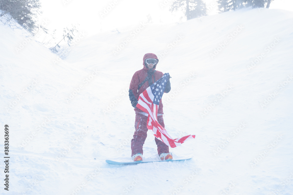 winter, leisure, sport and people concept - snowboarder sits high in the mountains on the edge of the slope and looks into the distance. snowboarder holding american flag