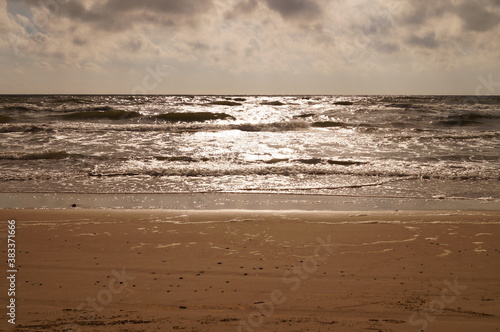 Sunset at the baltica beach, sun behind clouds reflects on water and waves with foam hitting sand. photo