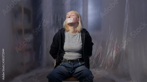 Female hostage sitting on a chair in a room with her mouth taped shut