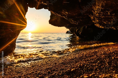 A small cave by the sea during sunset. Bright rays of the sun shine into the cave. Focus on pebbles and water's edge.