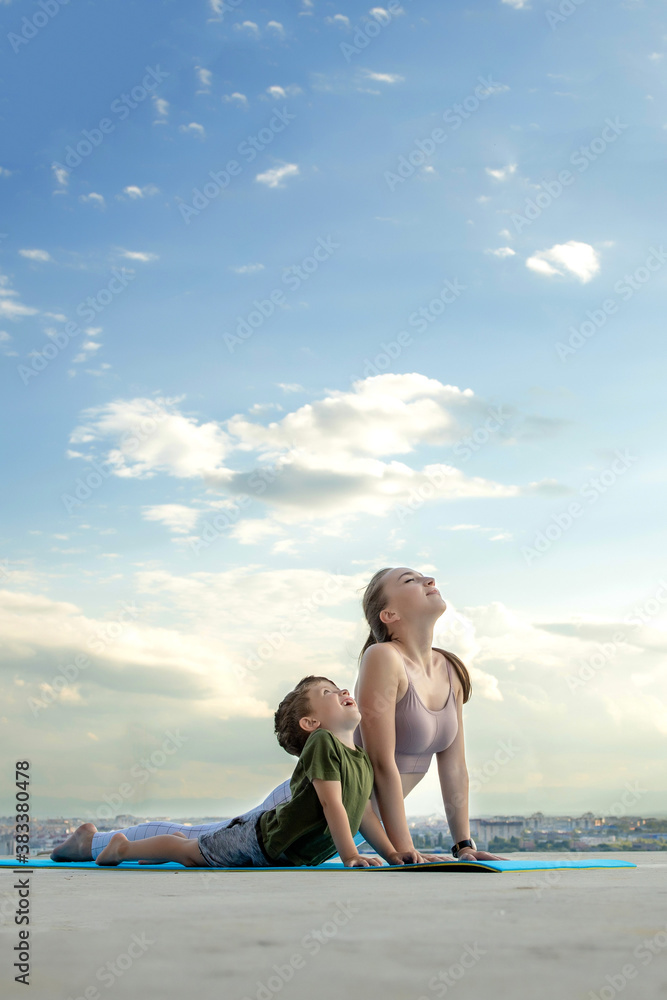 Mother and son doing exercise on the balcony in the background of a city during sunrise or sunset, concept of a healthy lifestyle