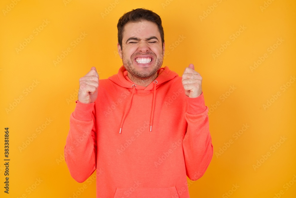 Handsome man with sweatshirt over isolated yellow background being excited for success with raised arms and closed eyes celebrating victory. Winner concept. Handsome man with sweatshirt over isolated yellow background being excited for success with raised arms and closed eyes celebrating victory. Winner concept.