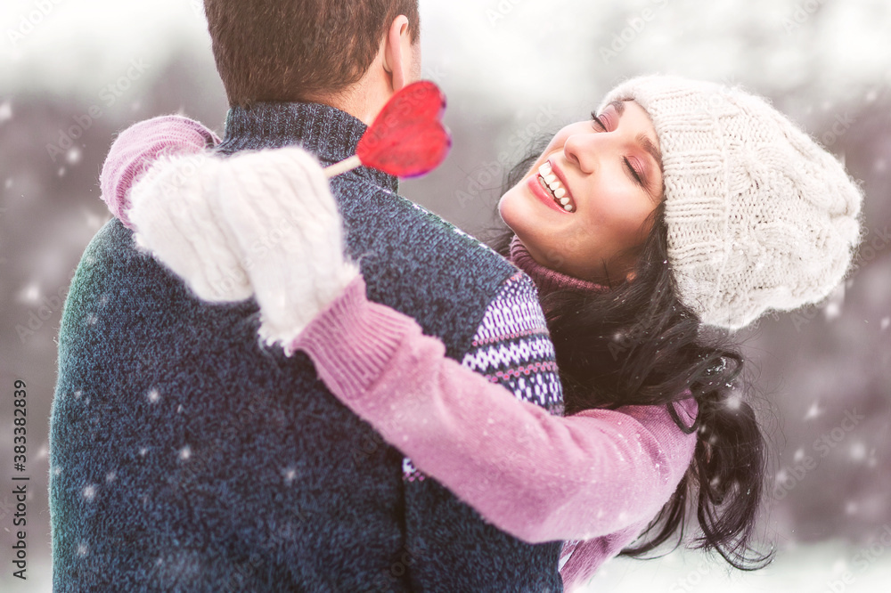 Outdoors romantic portrait of happy smiling couple in love. Holding in their hands red lollipop in shape of heart. Wearing knitted hat, mittens and sweaters. Love concept. Valentine day. It's snowing Outdoors romantic portrait of happy smiling couple in love. Holding in their hands red lollipop in shape of heart. Wearing knitted hat, mittens and sweaters. Love concept. Valentine day. It's snowing