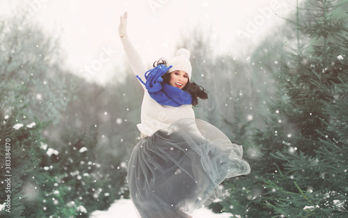 Winter fashion lifestyle portrait of charming young woman enjoying snowfall in snowy park. Smiling  jumping carefree  having fun. Wearing long tulle skirt  blue scarf. It s snowing. Happiness concept.