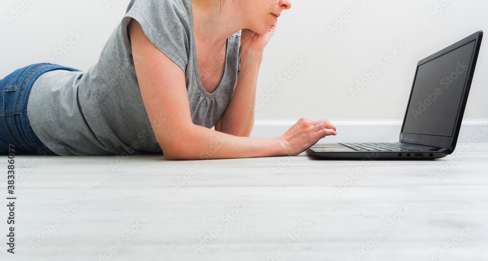 Fototapeta premium Croped caucasian woman lying on floor and working with a laptop. Female in casual clothes with t-shirt and jeans relax at home with internet. Copy space. Banner