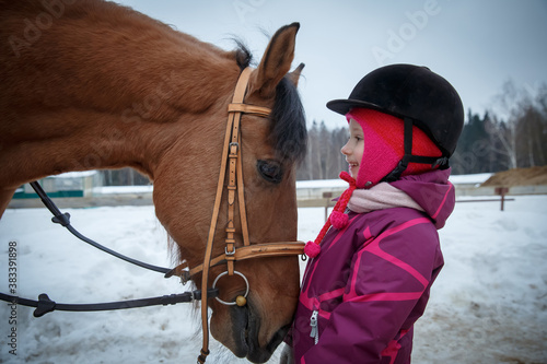 Close up of a little girl jockey smiling and looking to the horse eyes. Girl horse rider with a horse in a winter open riding hall, looking eye to eye. Girl and horse friendship, equestrian sport