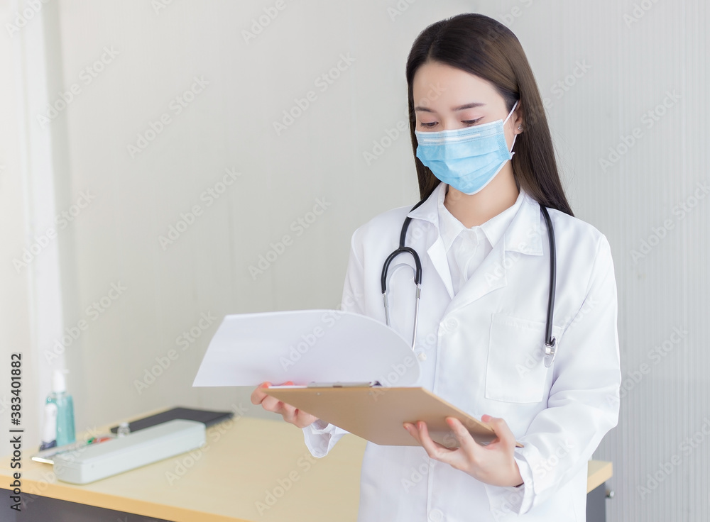 Asian woman doctor work and look at the report of patient in hospital. She wears a medical mask in Coronavirus pandemic situation.