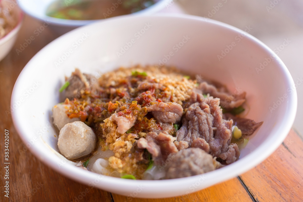 Meatballs and meat with wide rice noodles in soup was added pickled chili on top in a white bowl  on woody background. It is a Asian traditional food in Thailand.