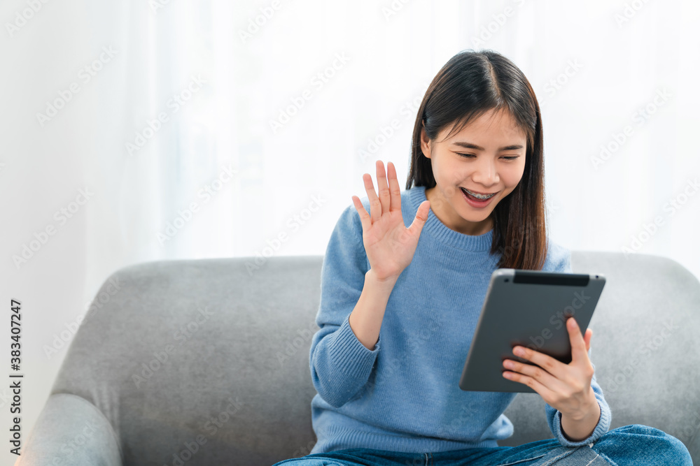Cheerful beautiful Asian woman in a blue shirt making video call online talking with friends and hand touching camera on digital tablet at sofa. Cheerful beautiful Asian woman in a blue shirt making video call online talking with friends and hand touching camera on digital tablet at sofa.