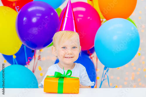 A little girl is sitting in a festive hat on a background of colorful balloons next to a gift and looks at the camera with a smile. First birthday concept