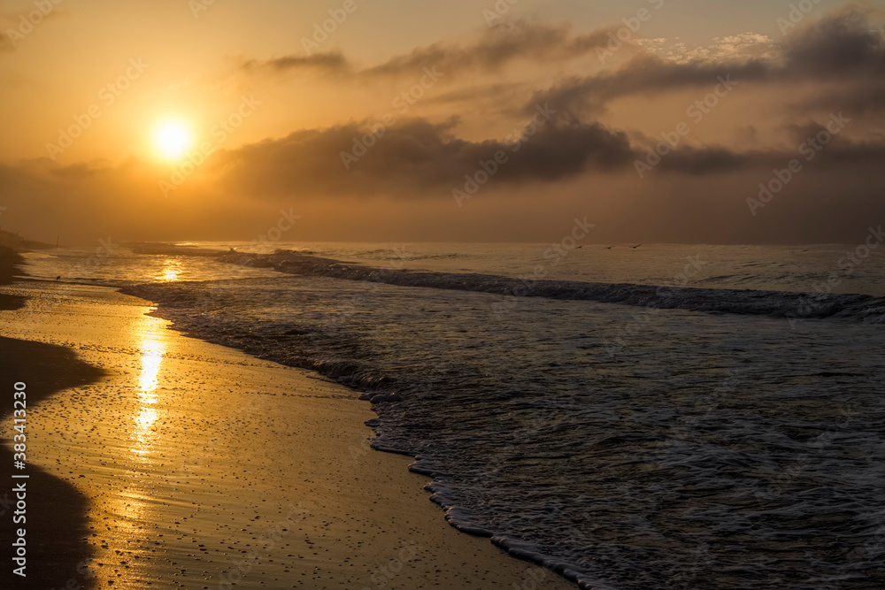 The golden morning light of sunrise shines on coastal Gulf Shores, Alabama with ocean waves gently breaking on the sandy beach and sea.