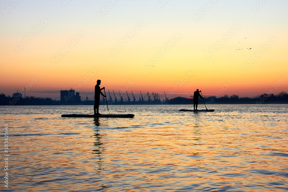 Silhouettes of two men paddle on stand up paddle boarding (SUP) on quiet winter or autumn Danube river at sunrise. Colorful sunset over the river with silhouettes of people