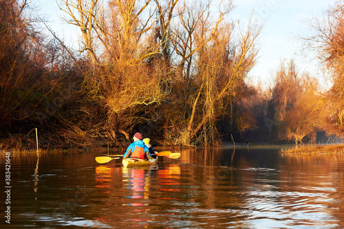 Kayaking in a calm winter morning at sunrise at the Danube river. A man and a woman row in yellow two-person kayak near shore in the rays of the rising sun