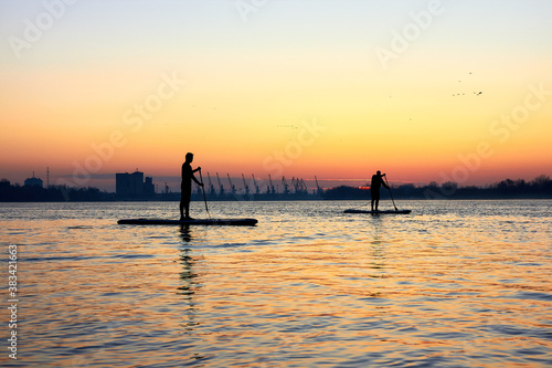 Silhouettes of two men paddle on stand up paddle boarding (SUP) on quiet winter or autumn Danube river at sunrise. Colorful sunset over the river with silhouettes of people