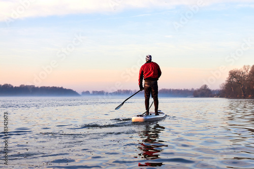Back view on man rowing on SUP (stand up paddle board) in autumn Danube river at foggy morning against the backdrop of autumn trees without leaves