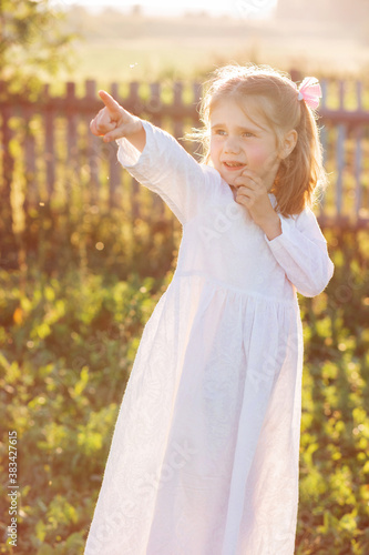 Little girl in a long white dress stands by the fence at sunset