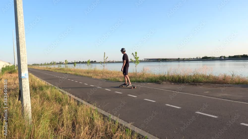 Hipster man riding longboard enjoying cruising on road near river bank in sunset light, wearing hat, sunglasses. Summer leisure activity, work out outside Hipster man riding longboard enjoying cruising on road near river bank in sunset light, wearing hat, sunglasses. Summer leisure activity, work out outside