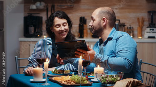 Young caucasian husband using tablet during festive meal showing to his wife holiday destinations. Adults sitting at the table  browsing online  surfing  using smartphones  enjoying the meal