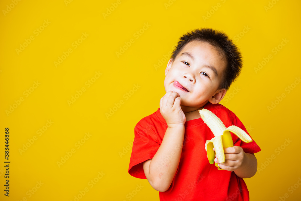 Happy portrait Asian child or kid cute little boy attractive smile wearing red t-shirt playing holds peeled banana for eating, studio shot isolated on yellow background Happy portrait Asian child or kid cute little boy attractive smile wearing red t-shirt playing holds peeled banana for eating, studio shot isolated on yellow background