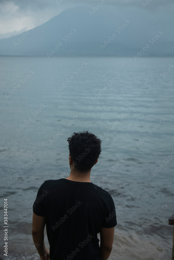 Young dark-haired man standing on the shore of the lake appreciating the landscape in front of him - Lake Atitlan in Guatemala on a cloudy day Young dark-haired man standing on the shore of the lake appreciating the landscape in front of him - Lake Atitlan in Guatemala on a cloudy day