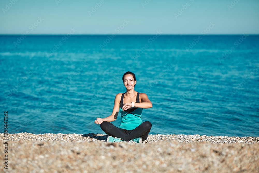 Woman sitting on beach and looking on watch. Calm sea and nature. Take care of health. Sport and active lifestyle idea