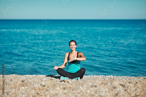 Woman sitting on beach and looking on watch. Calm sea and nature. Take care of health. Sport and active lifestyle idea