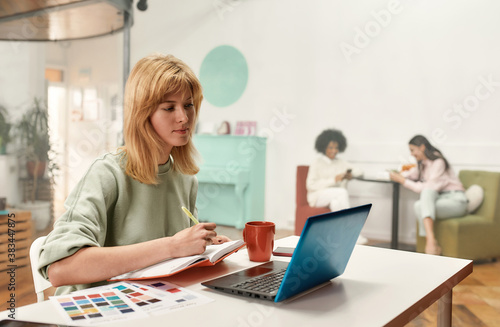 A creative young woman with blond hair sitting and working on her laptop while her colleagues sitting together on a background