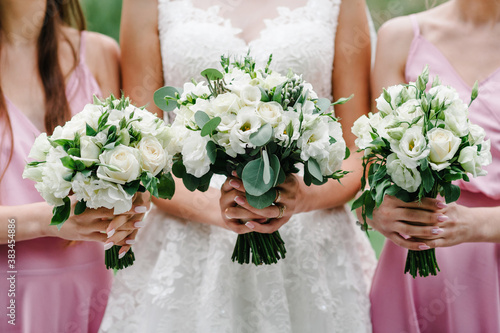 The bride and bridesmaids in an elegant pastel pink dress is standing and holding hand bouquets of flowers and greens at nature. Young beautiful girls holds a wedding bouquet outdoors.