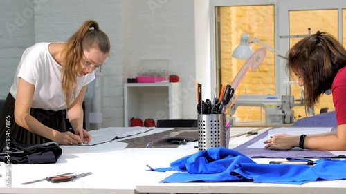 Two seamstresses sit at sewing machines, sew clothes. Female tailor is wearing a medical protective mask in a sewing workshop using sewing machine for tailoring clothing.
