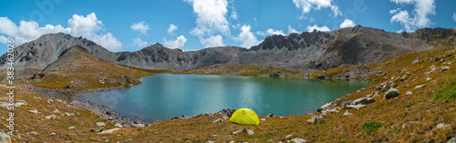 Panorama with a yellow tent on the shore of a mountain lake framed by rocky mountains. Kazakhstan, Kensu gorge