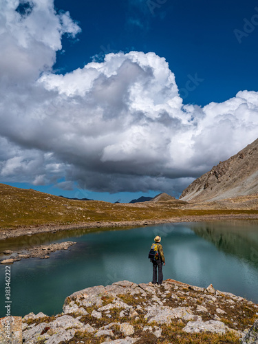 Male tourist near the shore of a high mountain lake. Large white clouds and mountains are reflected in the turquoise water. Kazakhstan, Kensu gorge