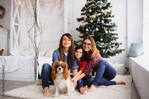 Two women and little girl with dog hugging near Christmas tree