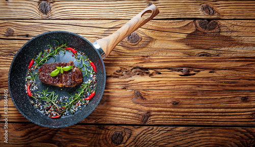 Black pan with white dots on an old wooden background. In the pan is grilled steak, chilli peppers, pepper, rosemary and salt.  Wooden background with copy space for text.