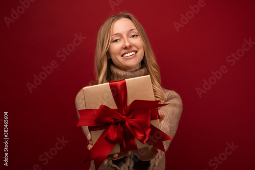 Charming blonde woman holding gift with red ribbon. Studio shot red background. New Year Birthday Holiday concept