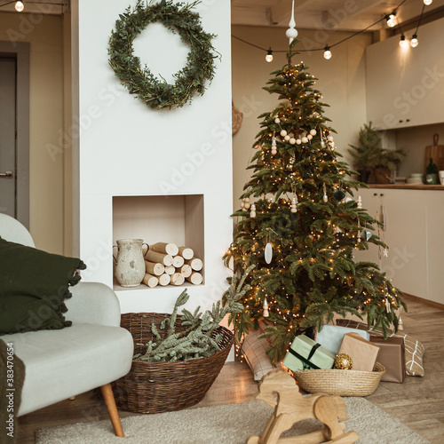 Modern home interior design concept. Comfortable cozy living room decorated with Christmas tree with gifts, wreath frame, fireplace, sofa, plaid, carpet. Christmas / New Year celebration decorations. photo