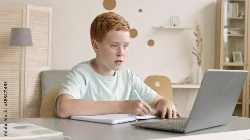 Pan shot of Caucasian school boy with ginger hair sitting at desktop in front of laptop at home and doing homework Pan shot of Caucasian school boy with ginger hair sitting at desktop in front of laptop at home and doing homework