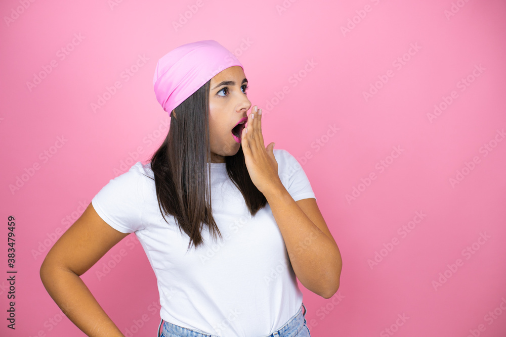 Young beautiful woman wearing pink headscarf over isolated pink background with her hands over her mouth and surprised, looking side Young beautiful woman wearing pink headscarf over isolated pink background with her hands over her mouth and surprised, looking side