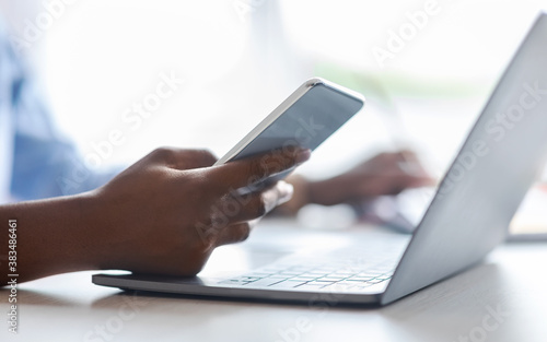 Unrecognizable black woman using smartphone and working on laptop in office, closeup