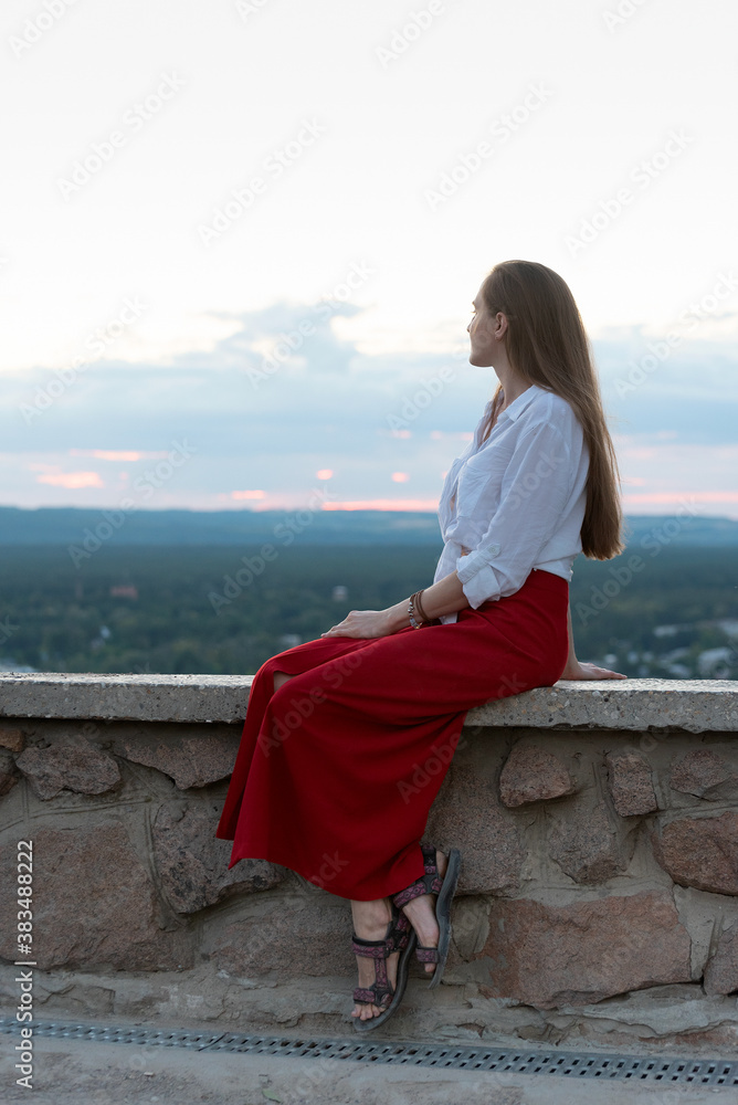 Young woman in red skirt sits on observation deck, nature on background. Vertical frame