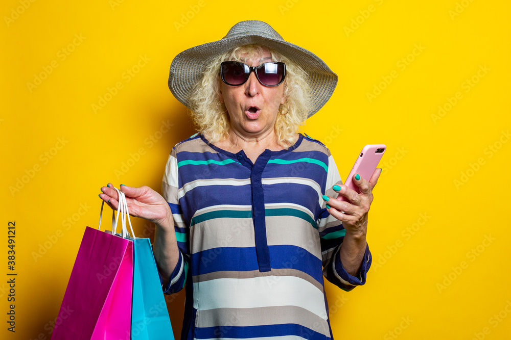 Old woman with surprised face in hat and glasses holds shopping bags with purchases and phone on yellow background Old woman with surprised face in hat and glasses holds shopping bags with purchases and phone on yellow background