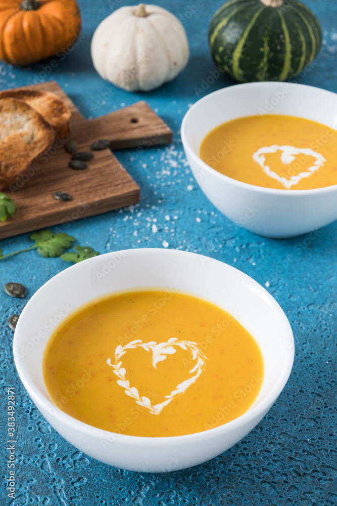 Two bowls of pumpkin soup with cream on blue background, decorated with little pumpkins, garlic and bread. Vertical stock photo. Two bowls of pumpkin soup with cream on blue background, decorated with little pumpkins, garlic and bread. Vertical stock photo.