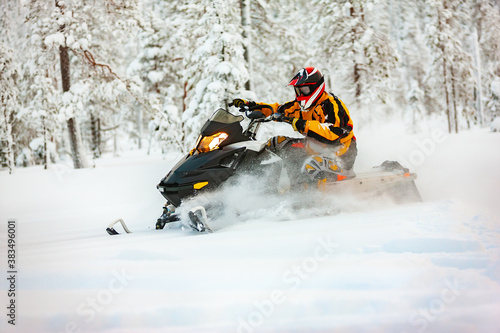 The racer in the outfit of a yellow-black overalls and a red-black helmet, driving a snowmobile at high speed riding through deep snow against the background of a snowy forest.