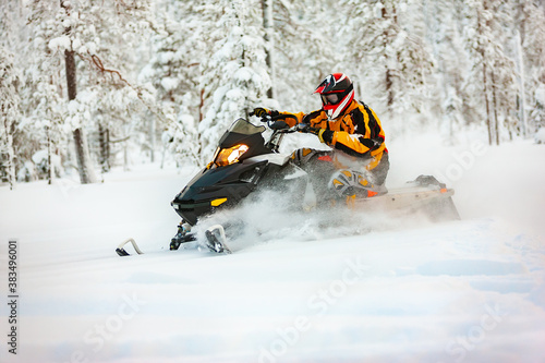 The racer in the outfit of a yellow-black overalls and a red-black helmet, driving a snowmobile at high speed riding through deep snow against the background of a snowy forest. photo