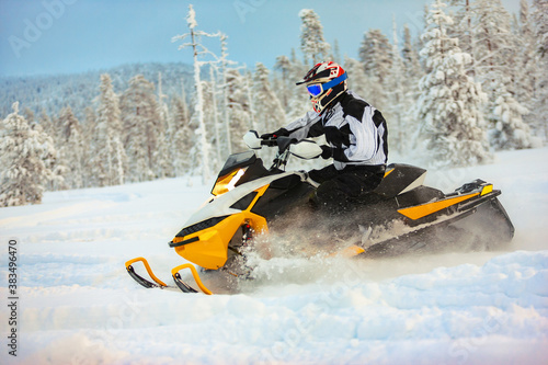 The rider in gear with a helmet drifting on a snowmobile on a deep snow surface on a background of snowy landscaping nature and winter forest.