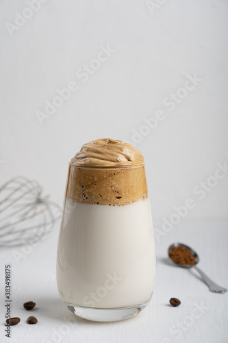 Single glass of popular korean dalgona drink made by whipping instant coffee powder, sugar and hot water with addition of cold milk on white wooden background with kitchen utensil. Vrtical orientation photo