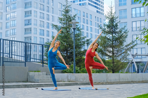 Young attractive woman in red and blue sportswear practices yoga exercises outdoors.