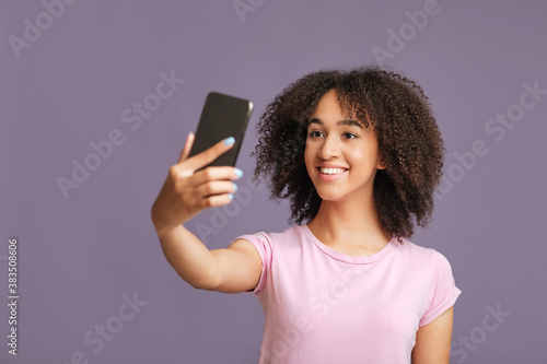 Modern technology, mobile app and selfie for fashion blog. Smiling african american woman makes photo on smartphone isolated on purple background