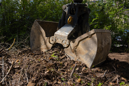 Heavy industrial machine scoop wooden branches and dirt of the ground. Hydraulic shovel lifting wood and waste from the garden
